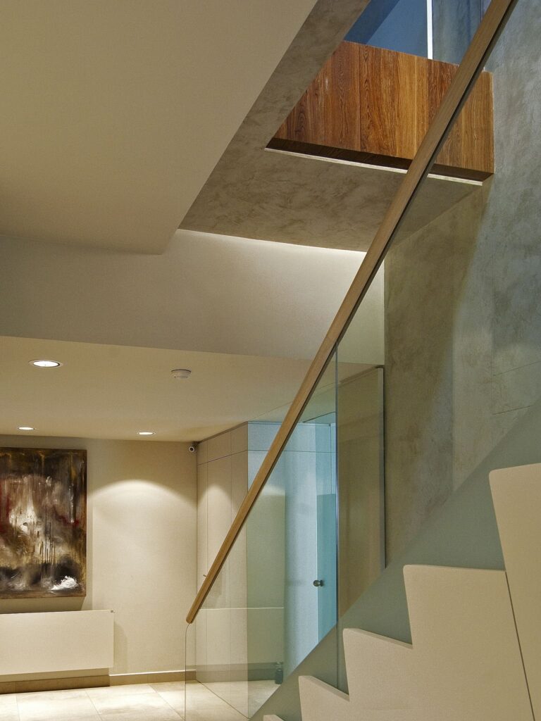 Escalera Interior Moderna y Elegante con Barandilla de Vidrio e Iluminación Natural Una escalera interior contemporánea que presenta escalones blancos elegantes y una barandilla de vidrio, iluminada por la luz natural que entra por una ventana. La escalera conduce a un nivel superior, mostrando un diseño minimalista con tonos neutros, incluyendo paredes grises y beiges, complementadas por obras de arte modernas.