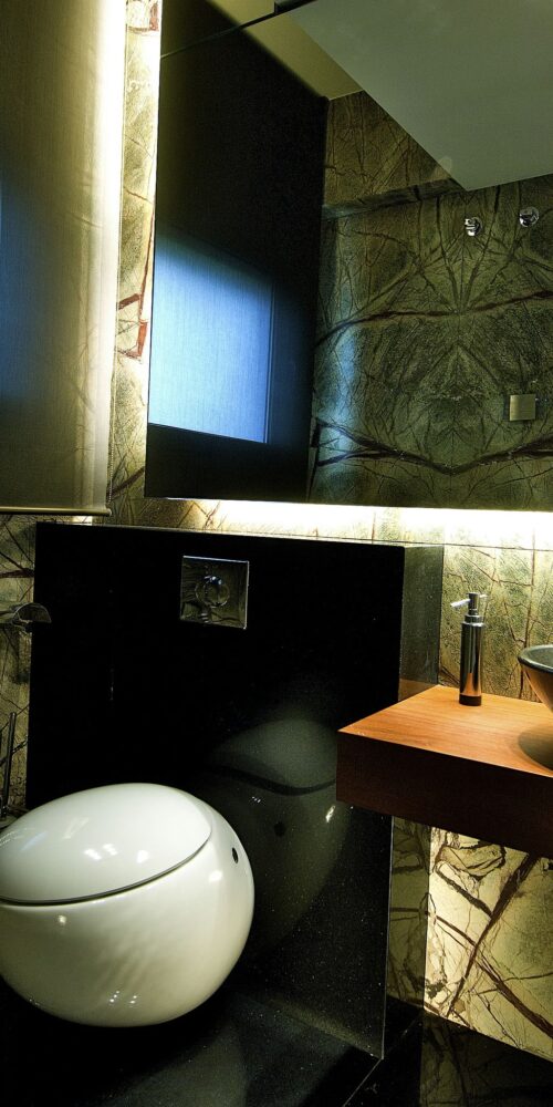Un baño contemporáneo que cuenta con un inodoro redondo blanco elegante, un lavabo negro estiloso sobre una encimera de madera, paredes de piedra texturizada, iluminación ambiental suave y un panel espejo que refleja elementos de diseño minimalista, complementado por un radiador de toallas montado.