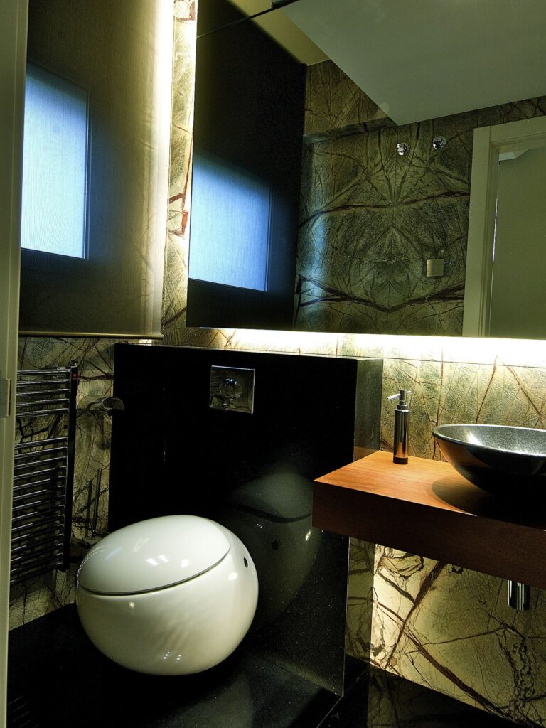 Baño Moderno con Inodoro Redondo Elegante y Lavabo Negro Estiloso Un baño contemporáneo que cuenta con un inodoro redondo blanco elegante, un lavabo negro estiloso sobre una encimera de madera, paredes de piedra texturizada, iluminación ambiental suave y un panel espejo que refleja elementos de diseño minimalista, complementado por un radiador de toallas montado.