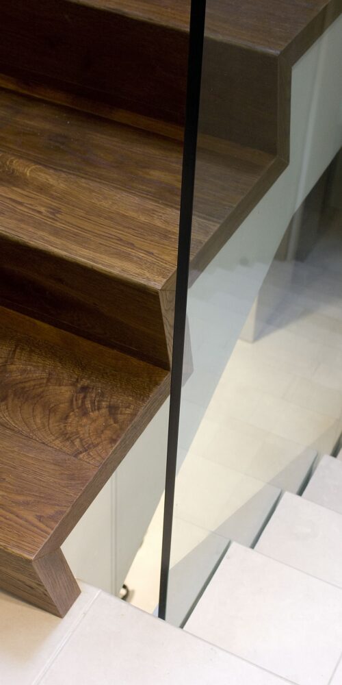 Una escalera moderna que presenta ricos escalones de madera texturizada con bordes angulares, complementada por una elegante barandilla de vidrio transparente. La escalera se encuentra sobre un suelo de baldosas de color claro, mostrando una sofisticada combinación de materiales naturales y elementos de diseño contemporáneo.
