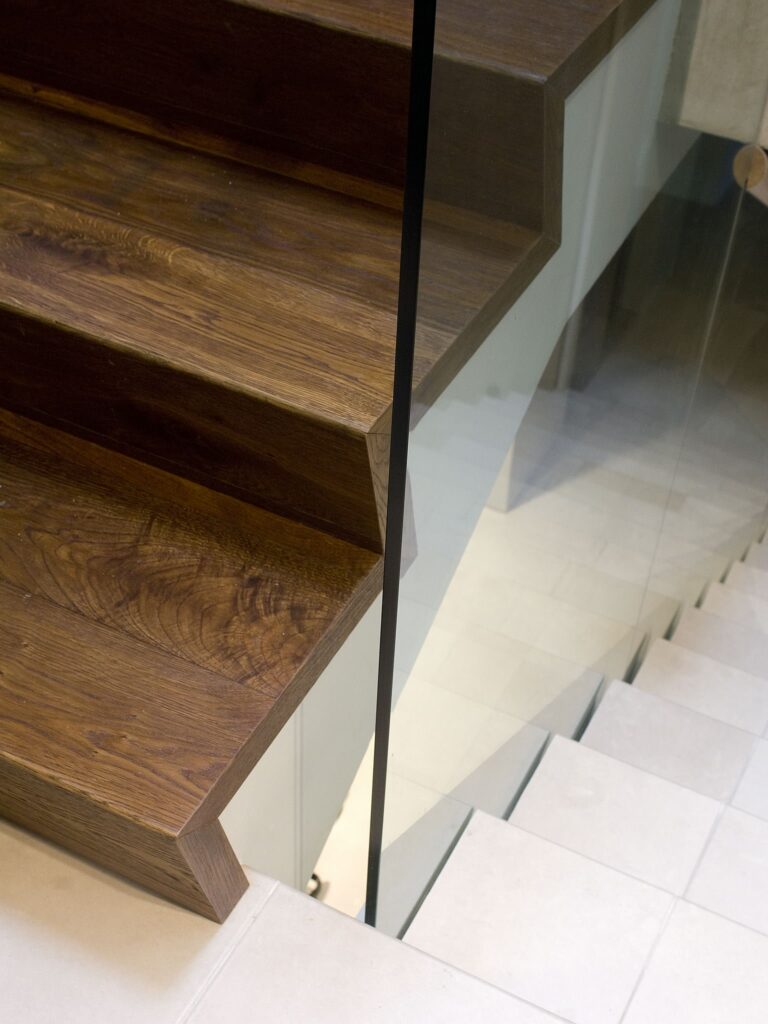 Escalera de Madera Moderna con Barandilla de Vidrio en Diseño Interior Contemporáneo Una escalera moderna que presenta ricos escalones de madera texturizada con bordes angulares, complementada por una elegante barandilla de vidrio transparente. La escalera se encuentra sobre un suelo de baldosas de color claro, mostrando una sofisticada combinación de materiales naturales y elementos de diseño contemporáneo.