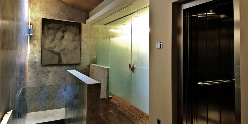 Un interior contemporáneo que muestra una escalera que lleva a un nivel superior, con una pared de vidrio, piso de madera, un ascensor a la derecha y arte abstracto en la pared, iluminado por una luz tenue.