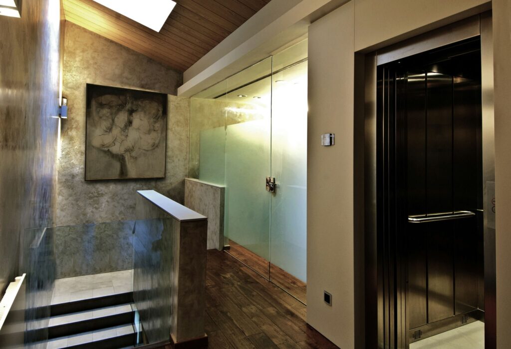 Interior Moderno Elegante con Pared de Vidrio y Arte Abstracto Un interior contemporáneo que muestra una escalera que lleva a un nivel superior, con una pared de vidrio, piso de madera, un ascensor a la derecha y arte abstracto en la pared, iluminado por una luz tenue.