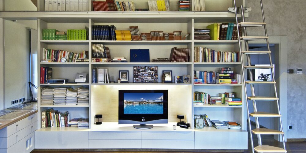 Una sala de estar contemporánea que muestra una estantería alta llena de libros y elementos decorativos, acompañada de una elegante unidad de entretenimiento con un televisor de pantalla plana. Una escalera rodante está posicionada contra la estantería, mejorando el acceso a los estantes superiores. El suelo de madera y la iluminación suave crean una atmósfera acogedora.