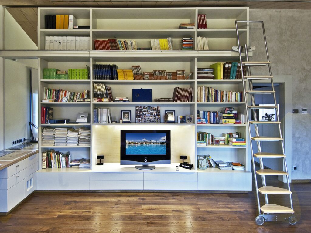 Espacio de Vida Moderno con Estantería Alta y Unidad de Entretenimiento Una sala de estar contemporánea que muestra una estantería alta llena de libros y elementos decorativos, acompañada de una elegante unidad de entretenimiento con un televisor de pantalla plana. Una escalera rodante está posicionada contra la estantería, mejorando el acceso a los estantes superiores. El suelo de madera y la iluminación suave crean una atmósfera acogedora.