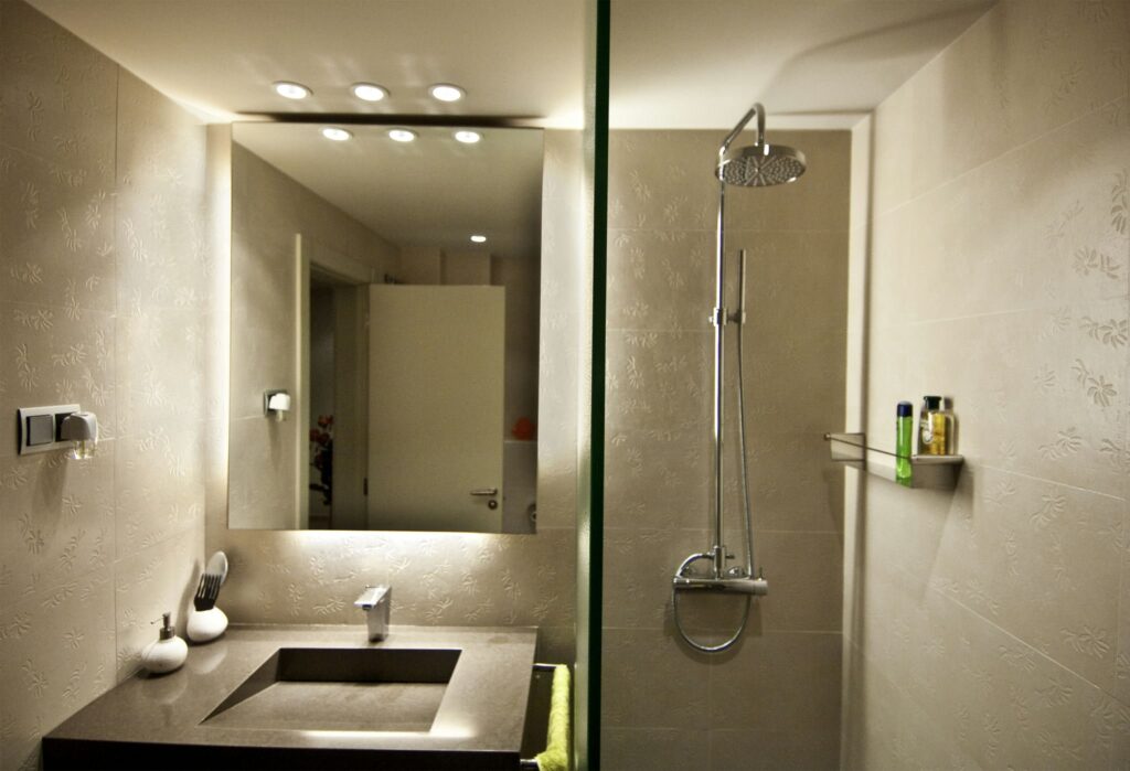 Diseño de Baño Moderno y Minimalista con Lavabo Cuadrado y Grifo Elegante Un baño moderno y elegante que presenta un lavabo cuadrado combinado con un grifo elegante sobre una encimera oscura, realzado por iluminación empotrada. La pared cuenta con delicados patrones florales mientras que a la derecha se encuentra una mampara de ducha de vidrio. Una estantería estrecha que sostiene una variedad de artículos de tocador añade a la estética contemporánea, todo presentado en tonos neutros para transmitir un aspecto limpio y minimalista.