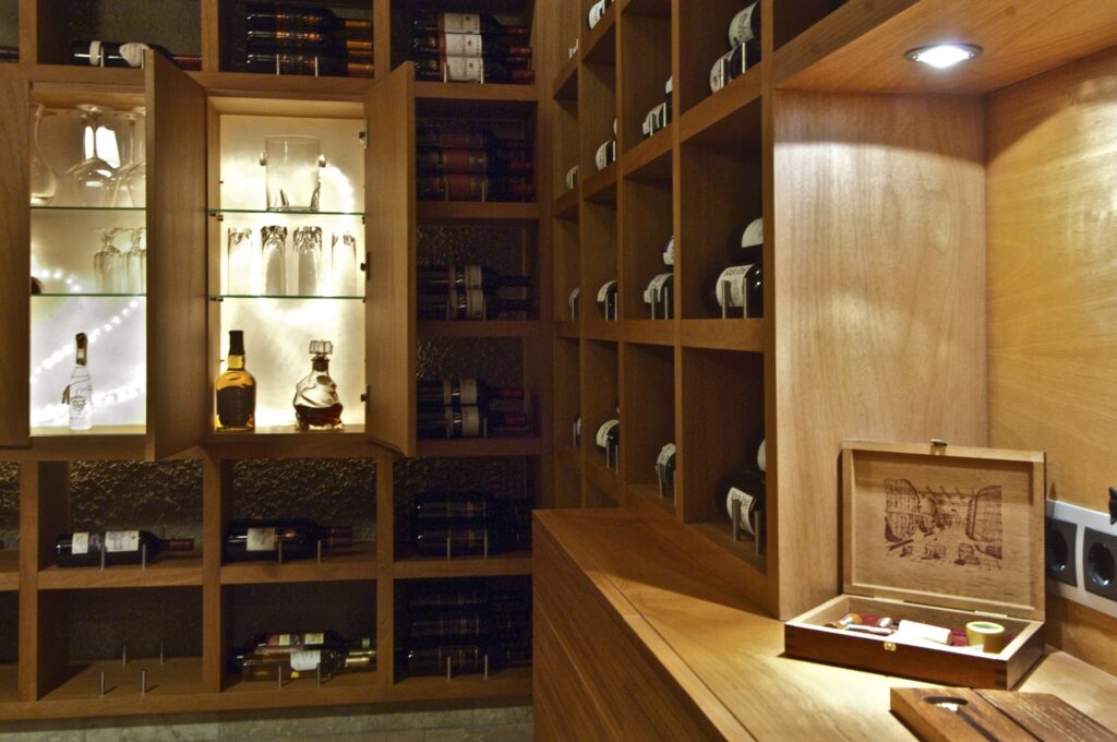 Bodega Elegante con Muebles de Madera y Iluminación Ambiental Bodega elegante con mobiliario de madera, estantes iluminados llenos de botellas de vino, un armario de vidrio para licores y cristalería, y una caja de madera decorativa, todo realzado por una iluminación suave.