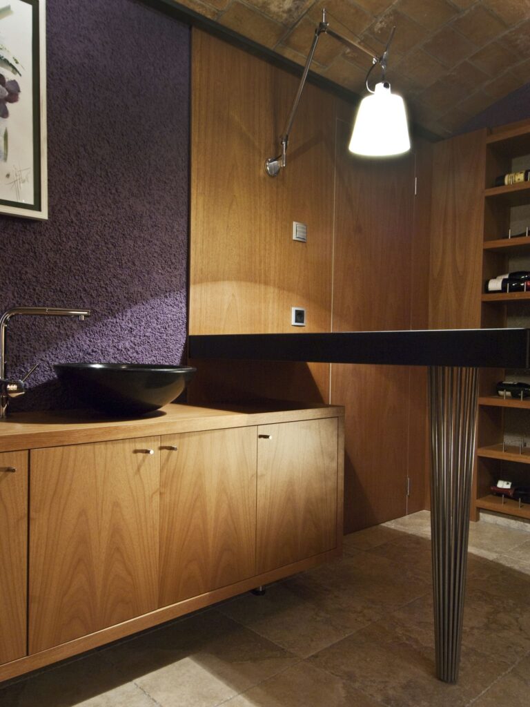 Interiores de baño modernos y minimalistas con encimera oscura y acentos vibrantes Un interior de baño contemporáneo que presenta una elegante encimera oscura sobre patas únicas de metal con nervaduras verticales, un lavabo redondo negro y un elegante armario de madera, resaltado por paredes texturizadas de color púrpura y suave iluminación colgante.