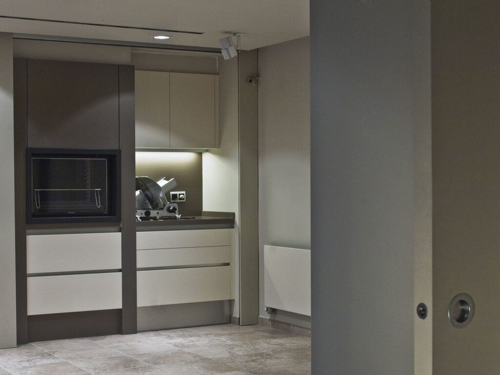 Diseño de Cocina Moderna Minimalista con Muebles Elegantes y Luz Suave Una cocina moderna bellamente diseñada que presenta una estética minimalista, exhibiendo muebles de tonos neutros, un horno empotrado, electrodomésticos de encimera elegantes y un espacioso suelo de baldosas. La luz suave realza la elegancia de los muebles, mientras que una pared parcial insinúa un diseño de planta abierta más amplio más allá de la vista.