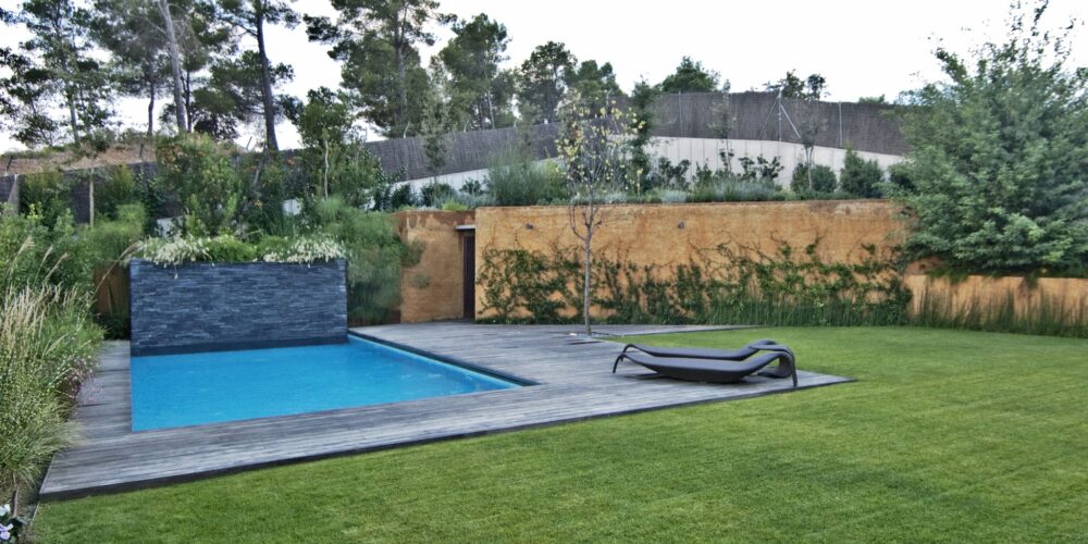 Un espacio exterior moderno que presenta una serena piscina rodeada por una terraza de madera, complementada por una silla de descanso y una pared de piedra adornada con vegetación, enmarcado por altos árboles que crean una atmósfera de paz.