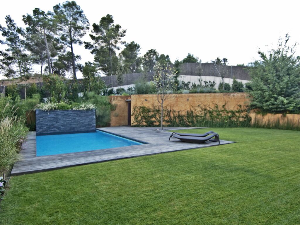 Tranquila Piscina Moderna al Aire Libre Rodeada de Vegetación Exuberante Un espacio exterior moderno que presenta una serena piscina rodeada por una terraza de madera, complementada por una silla de descanso y una pared de piedra adornada con vegetación, enmarcado por altos árboles que crean una atmósfera de paz.