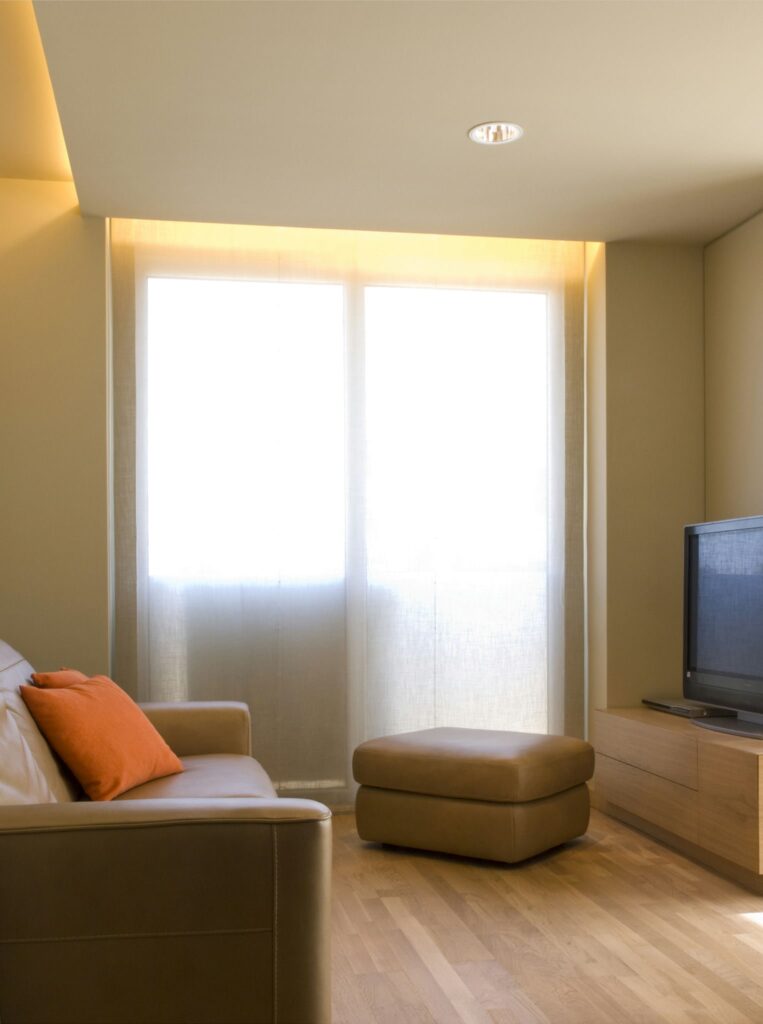 Diseño de Sala de Estar Moderna con Luz Natural y Estética Minimalista Una sala de estar moderna, brillante y acogedora con grandes ventanas y cortinas translúcidas, un sofá de cuero beige con un cojín naranja y un suelo de madera pulida, que ejemplifica un estilo de decoración minimalista.