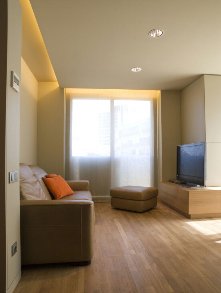 Sala de Estar Moderna y Elegante con Sofá de Cuero y Iluminación Cálida Una sala de estar moderna y luminosa que presenta un sofá de cuero color marrón claro adornado con un cojín decorativo naranja, un ottoman a juego, un televisor de pantalla plana montado en la pared junto a una consola de madera clara, y grandes ventanas con cortinas translúcidas que permiten que la luz natural ilumine el espacio.