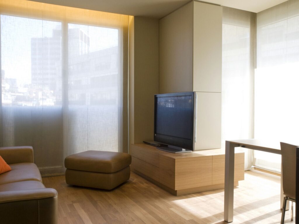 Diseño de interiores moderno en una vivienda de lujo en Barcelona Interior de un espacio moderno en Barcelona, con diseño contemporáneo, muebles de alta calidad y abundante luz natural. Destacan un sofá bajo, un ottoman, y una TV de pantalla plana montada en una customizada cabinetry de madera.