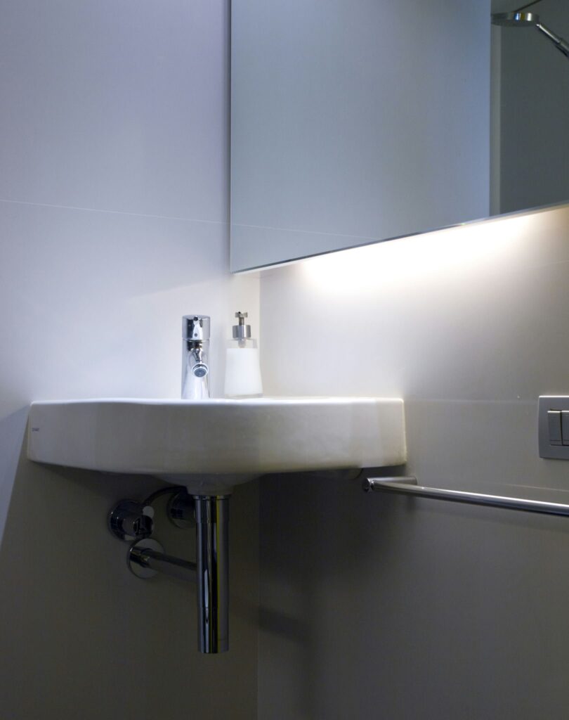 Baño Moderno Elegante con Lavabo Blanco y Accesorios de Cromo Baño moderno que presenta un lavabo blanco montado en la pared, grifo de cromo contemporáneo, dispensador de jabón transparente, iluminación ambiental y barra de toallas de cromo, todo contra una pared de color claro.