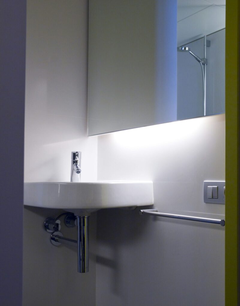 Esquina de baño minimalista moderno con lavabo blanco y grifo cromado Esquina de baño contemporáneo que muestra un lavabo blanco montado en la pared con un grifo cromado, iluminado por una tira de luz elegante. Las paredes de tonos neutros y el diseño minimalista crean una atmósfera limpia y acogedora. Un toallero y un interruptor añaden funcionalidad al espacio.
