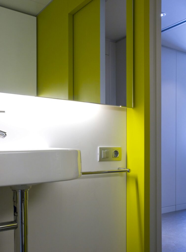 Interior de baño moderno y elegante con acentos coloridos Un baño moderno y estiloso que presenta un diseño minimalista con un lavabo elegante, paredes blancas y un acento amarillo vibrante. La iluminación suave crea una atmósfera limpia mientras que un toallero de cromo añade funcionalidad. La reflexión en el espejo revela espacio adicional a través de una puerta abierta, enfatizando la estética contemporánea en áreas compactas.