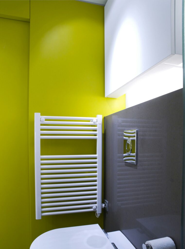 Interior de baño moderno vibrante con elementos de diseño elegantes Interior de baño moderno que muestra una pared verde lima brillante contrastada por una pared gris oscuro, con un toallero eléctrico blanco, armarios de almacenamiento ocultos y un lavabo blanco elegante en un espacio bien iluminado.