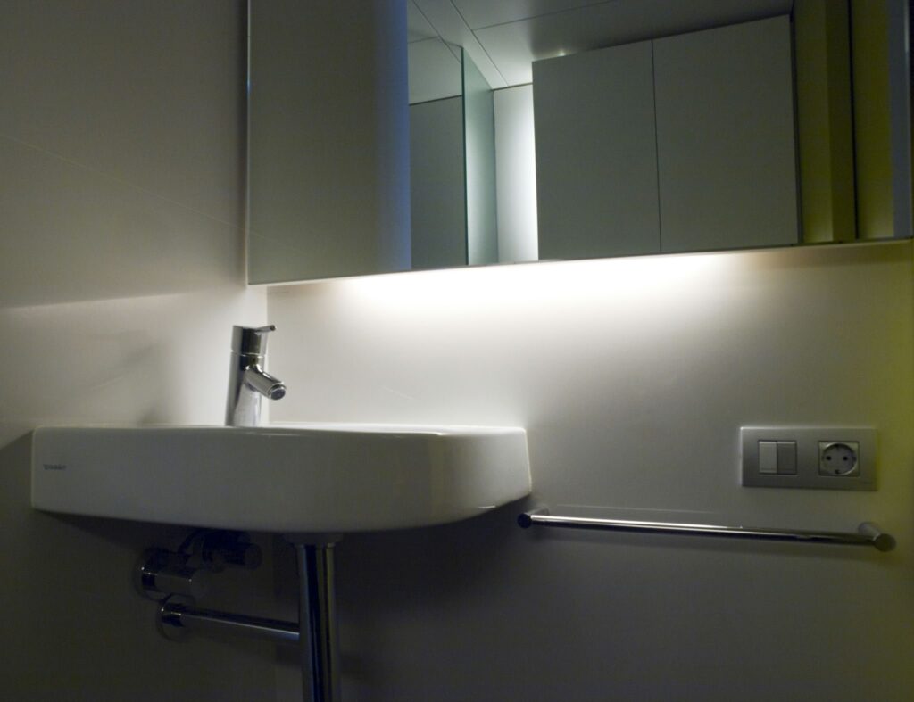 Diseño de Baño Minimalista Elegante con Lavabo Blanco Moderno y Grifo de Cromo Un baño moderno que presenta un diseño minimalista con un lavabo blanco elegante, grifo de cromo, reflejo de iluminación ambiental en el espejo y líneas limpias contra una pared de color claro.