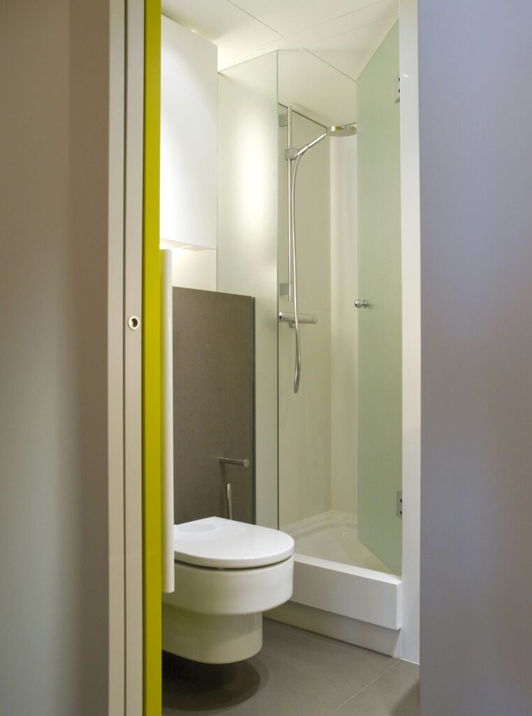 Baño Compacto Moderno con Ducha de Cristal y Acentos Verde Lima Un baño moderno y elegante que presenta un inodoro redondo blanco montado en la pared, un recinto de ducha de vidrio transparente y un suelo de baldosas beige. El espacio se ilumina con luz natural y se acentúa con mobiliario verde lima, creando un diseño limpio y contemporáneo para espacios pequeños.