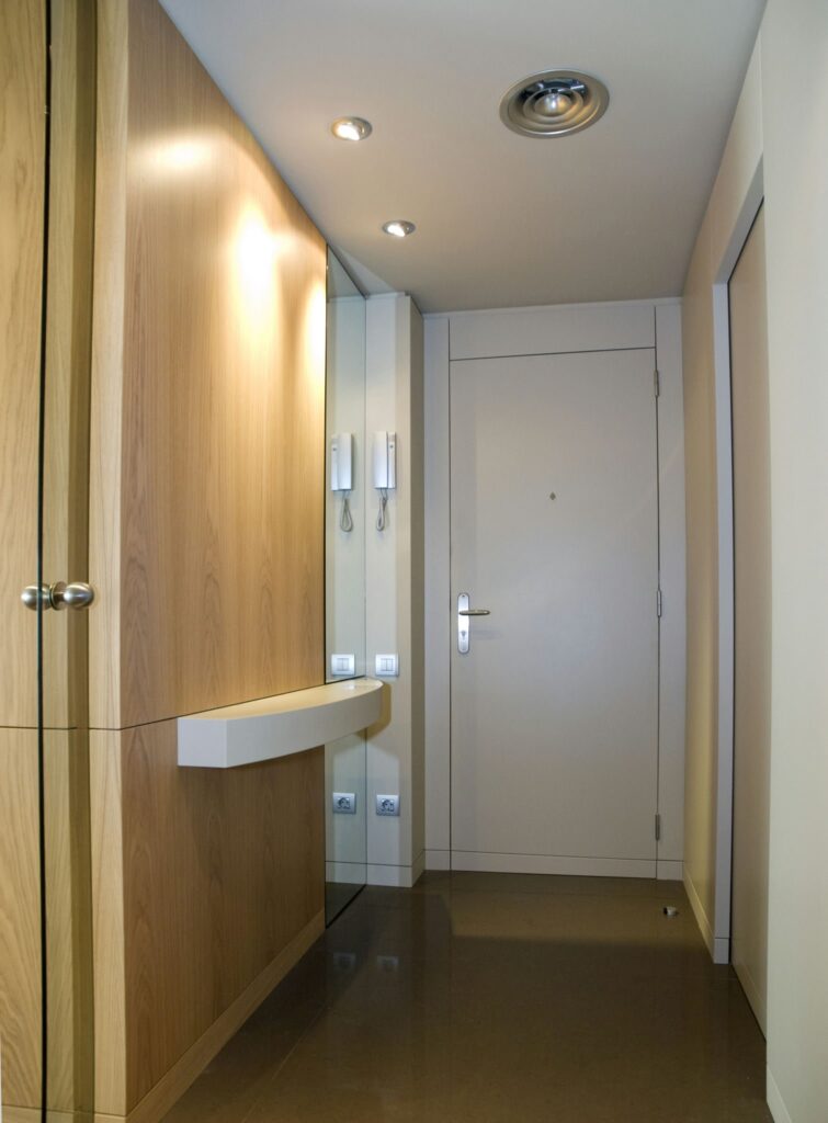 Entrada de Pasillo Moderna con Diseño Elegante y Acabados Pulidos Un pasillo moderno que muestra una unidad de almacenamiento de madera brillante, una puerta minimalista, un suelo pulido y videoporteros, todos contribuyendo a una atmósfera de entrada limpia y organizada.