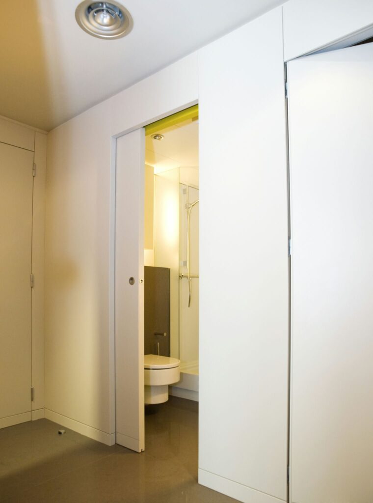Entrada de Baño Minimalista Contemporáneo con Diseño Elegante Entrada de baño moderno que muestra un diseño minimalista con una puerta parcialmente abierta que revela un interior luminoso, inodoro y un área de ducha elegante. Las paredes son de color claro y el suelo es suave en un tono neutro, realzando la estética contemporánea. Un accesorio de luz circular ilumina el espacio.