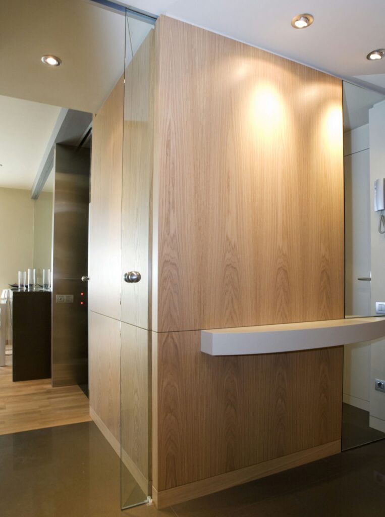 Diseño de interiores contemporáneo con elemento de pared de madera y elementos de vidrio Un espacio interior moderno que muestra una elegante pared de madera con una estantería horizontal, iluminada por una suave luz. Los elementos de vidrio de la pared crean un aspecto semitransparente. Adyacente a la pared hay un ascensor parcialmente visible en el lado izquierdo, mientras que el suelo reflectante y elegante realza la estética contemporánea de la habitación.