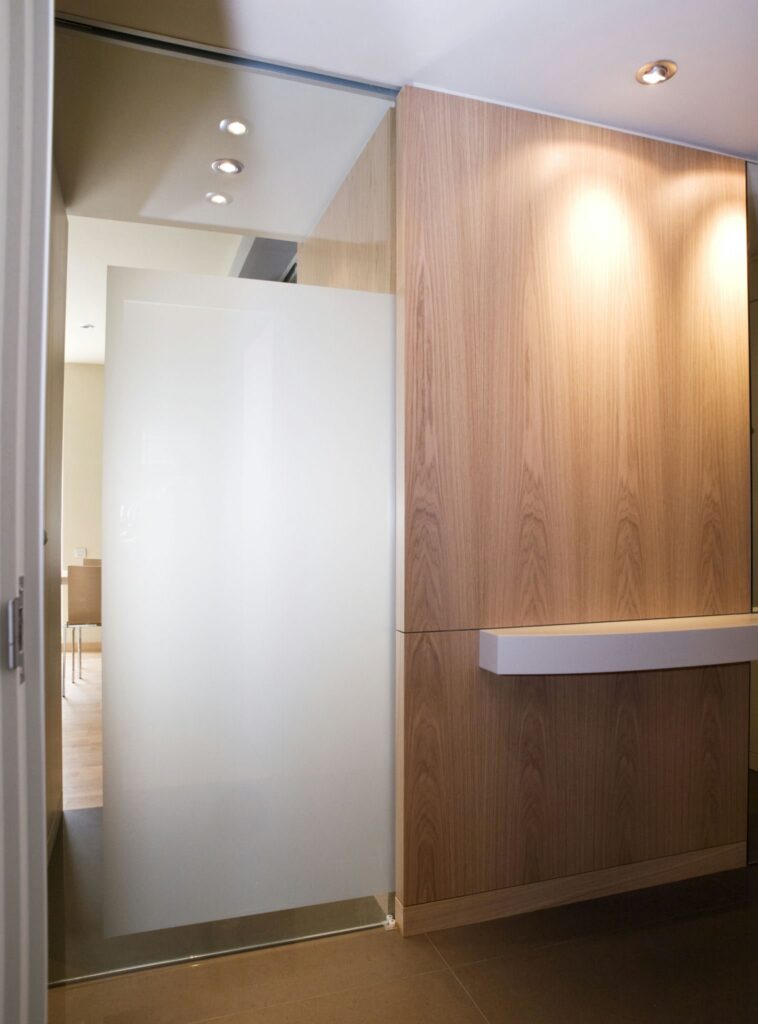 Diseño de Interior Moderno con una Puerta de Vidrio Esmerilado y Accentos de Madera Un espacio interior moderno, brillante y acogedor, que muestra una puerta de vidrio esmerilado parcialmente y una pared de madera. La puerta conduce a una habitación bien iluminada decorada con tonos neutros, iluminada por suaves luces de techo que realzan la cálida atmósfera. El suelo oscuro contrasta maravillosamente con los elementos más claros, encarnando principios de diseño contemporáneo centrados en el minimalismo y el uso de materiales naturales.