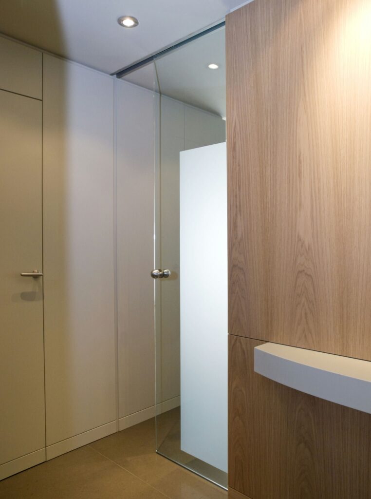 Interior de baño moderno con ducha de cristal y acentos de madera clara Vista interior de un baño moderno que muestra un elegante cerramiento de ducha de vidrio, gabinetes de madera clara y paredes en tonos neutros. La imagen destaca un mango elegante en un cajón de madera, acentuado por la iluminación empotrada que ilumina el espacio contemporáneo, ofreciendo una combinación de funcionalidad y atractivo estético.