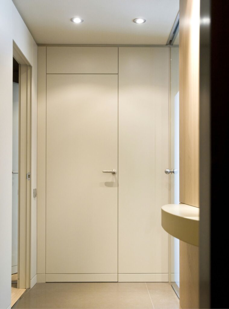 Diseño de Pasillo Interior Moderno y Minimalista Un pasillo interior contemporáneo y minimalista que presenta una pared beige con una puerta cerrada, luces empotradas en el techo para una iluminación brillante y una encimera redonda y suave. El suelo consiste en azulejos neutros que realzan la estética despejada, mostrando líneas limpias y una atmósfera brillante y aireada.