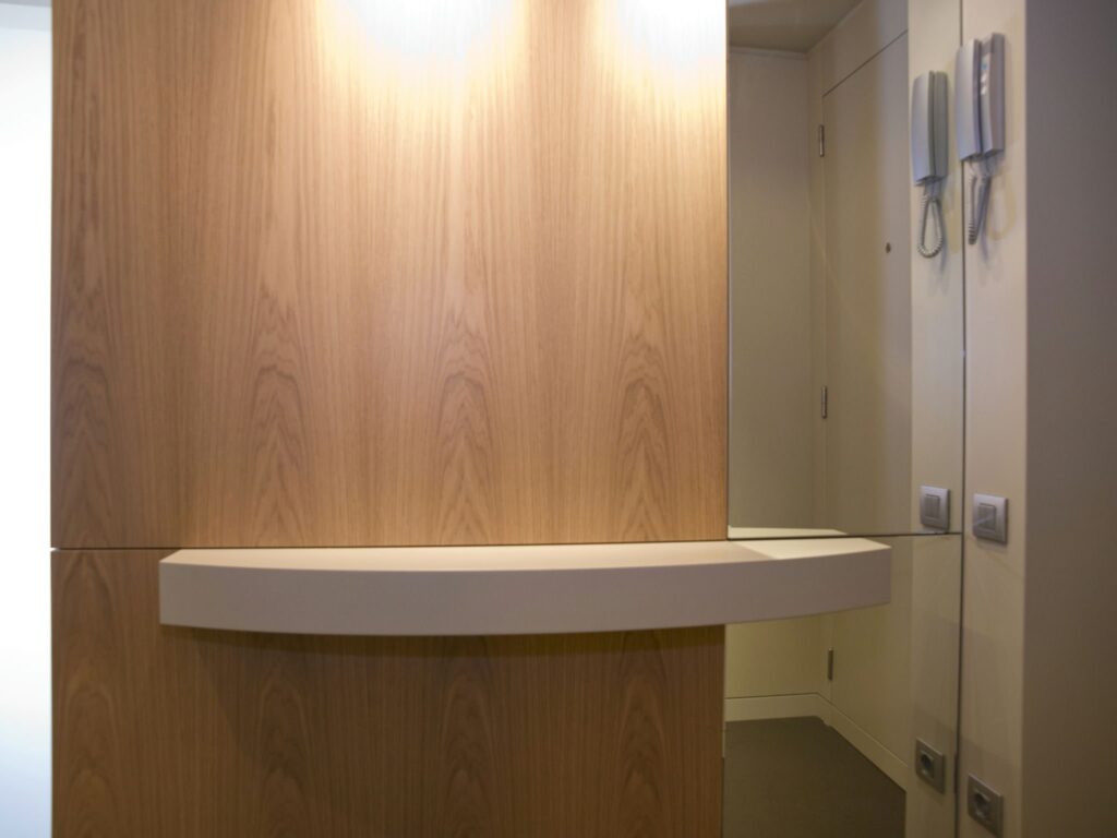 Entrada Contemporánea con Pared de Madera y Estante Curvado Un interior moderno que presenta una elegante pared de madera con un estante curvado iluminado por una suave iluminación, junto a una pared espejada equipada con dispositivos de intercomunicación, ejemplificando un diseño minimalista.