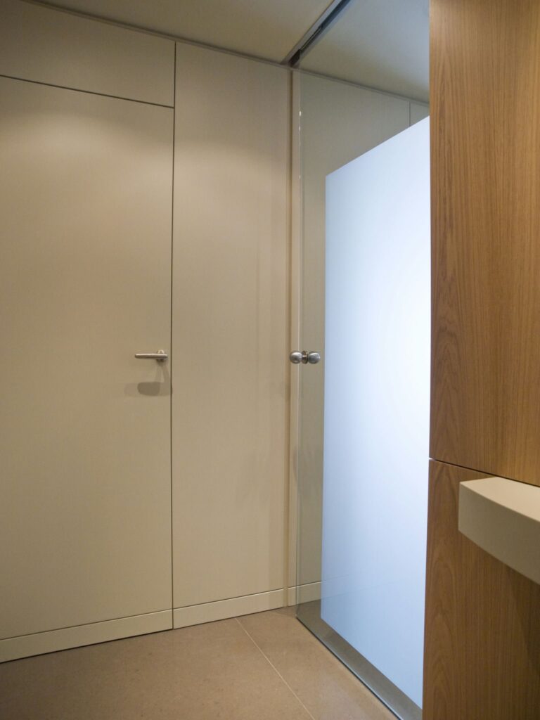 Espacio Interior Moderno Minimalista con Puerta de Color Claro y Panel de Vidrio Esmerilado Interior moderno con un diseño minimalista que presenta una puerta de color claro y un panel de vidrio esmerilado para privacidad. Una textura de pared de madera cálida agrega profundidad, contrastando con el suelo de baldosas de color claro.