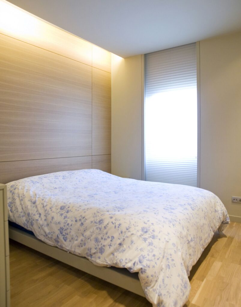 Interior de dormitorio sereno con edredón floral y luz natural Un dormitorio pacífico que muestra una cama bien hecha adornada con un suave edredón floral en tonos pastel, ubicada contra una pared de madera lisa, iluminada por la luz natural de una ventana brillante.
