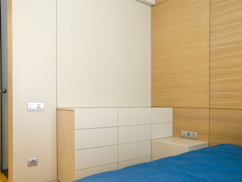 Esquina de Dormitorio Moderno con Paneles de Pared de Color Claro y Cómoda Elegante Una esquina de dormitorio moderno y sereno que presenta paneles de pared de color claro, una cómoda minimalista elegante y una colcha azul que añade un toque de color. El diseño resalta texturas suaves de blanco y madera, junto con interruptores de luz funcionales y tomacorrientes.