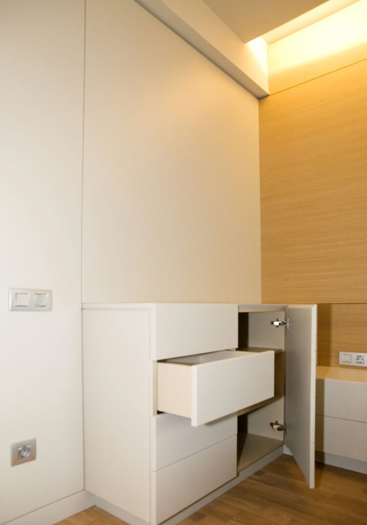 Diseño de armario moderno minimalista contra panel de madera cálido Un elegante armario moderno minimalista exhibido contra una pared clara, con una sección abierta y un solo cajón parcialmente expuesto. El armario es blanco con líneas limpias y está complementado por un panel de madera cálido en el costado. Las tomas de corriente son visibles junto al armario, y el suelo de madera clara realza el aspecto contemporáneo.