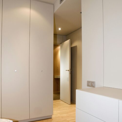 Un interior moderno y elegante que muestra muebles de color claro y suelo de madera. La imagen presenta una puerta parcialmente abierta que conduce a otra área del cuarto, enfatizando los elementos de diseño minimalista caracterizados por líneas limpias y tonos neutros. El espacio bien iluminado se realza con luces empotradas en el techo, creando una atmósfera acogedora, con una cómoda blanca en primer plano y un vistazo del espacio adyacente al fondo.