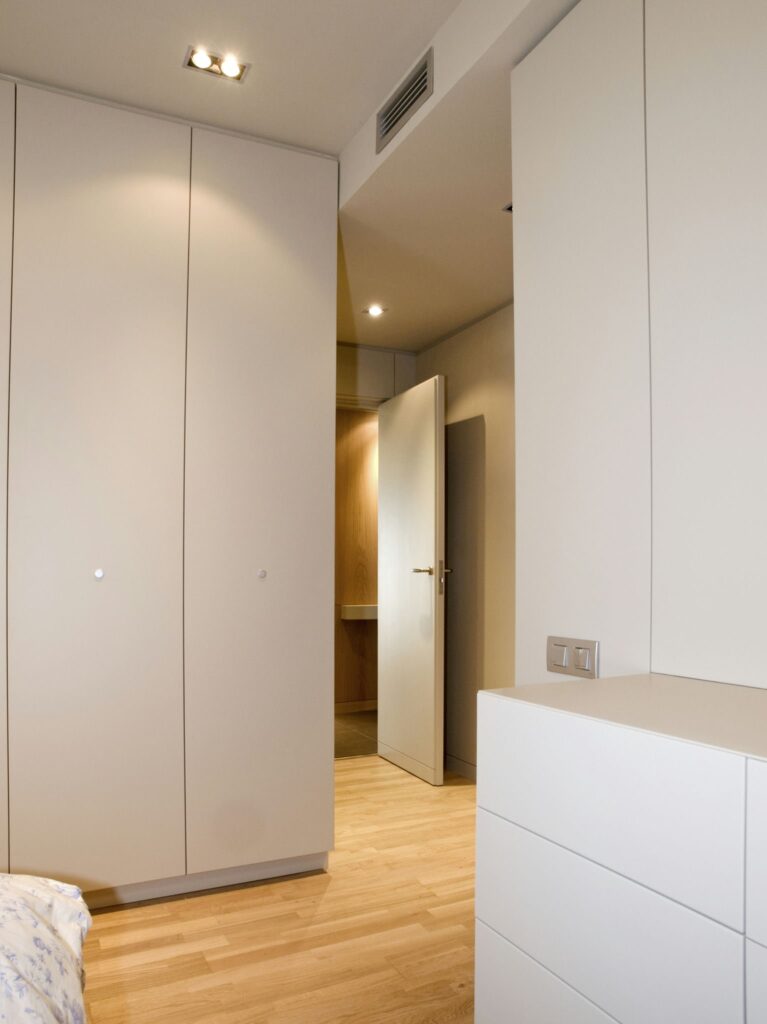 Interior Minimalista Moderno con Muebles de Color Claro y Suelo de Madera Un interior moderno y elegante que muestra muebles de color claro y suelo de madera. La imagen presenta una puerta parcialmente abierta que conduce a otra área del cuarto, enfatizando los elementos de diseño minimalista caracterizados por líneas limpias y tonos neutros. El espacio bien iluminado se realza con luces empotradas en el techo, creando una atmósfera acogedora, con una cómoda blanca en primer plano y un vistazo del espacio adyacente al fondo.