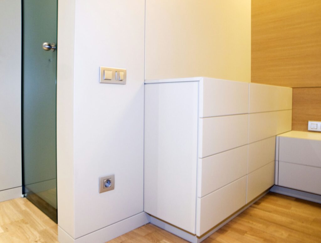 Interior Moderno Minimalista con Cofre de Almacenamiento Blanco y Acabados de Madera Claro Un rincón contemporáneo de un interior moderno que presenta un elegante cofre de almacenamiento blanco contra una pared de acabado de madera clara. Una puerta de vidrio entreabierta conduce a otra habitación, mostrando las líneas limpias y el espacio abierto del diseño minimalista. Los enchufes eléctricos y los interruptores de luz visibles añaden al estético moderno, enfatizando la funcionalidad y la simplicidad en la decoración del hogar.