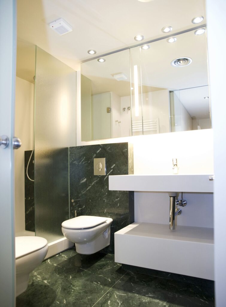 Diseño de baño moderno con mármol oscuro y accesorios elegantes Un baño moderno con un diseño elegante, que presenta un suelo de mármol oscuro, armarios blancos, un inodoro montado en la pared y un lavabo minimalista en una encimera espaciosa. Grandes espejos y focos de techo realzan la estética contemporánea y limpia, haciéndolo ideal para el diseño de interiores moderno.