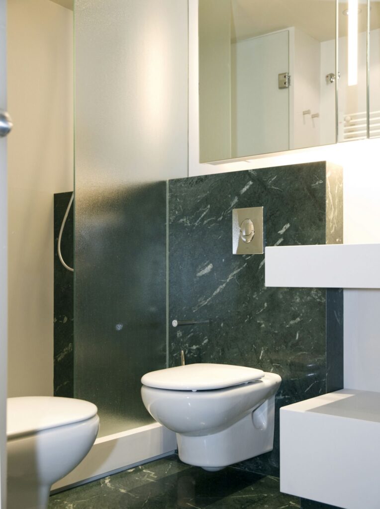 Baño moderno y lujoso con mampara de ducha de vidrio y acentos de mármol oscuro Interior de baño contemporáneo que muestra dos inodoros blancos, una mampara de ducha de vidrio elegante y paredes acentuadas con mármol oscuro. El diseño minimalista presenta un tocador blanco y un gran espejo que refleja el espacio estiloso, realzado por una iluminación suave para una atmósfera lujosa.