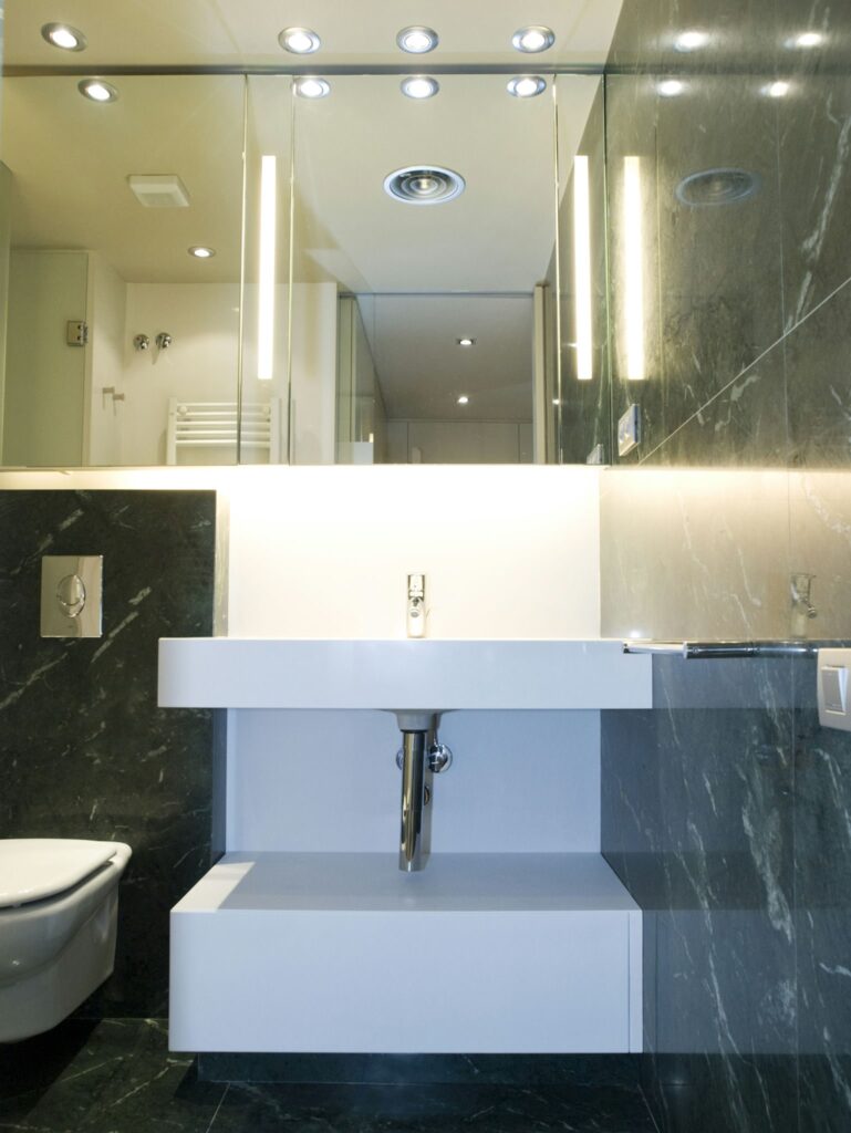 Baño Moderno de Lujo con Mármol Oscuro y Diseño Minimalista Un elegante baño moderno que muestra paredes y suelos de mármol oscuro con una unidad de lavabo blanca minimalista debajo de un gran espejo iluminado. El espacio cuenta con accesorios contemporáneos y un grifo de cromo pulido, añadiendo un toque sofisticado. Un inodoro compacto está posicionado contra la pared, enfatizando la estética limpia y funcional de este lujoso baño.