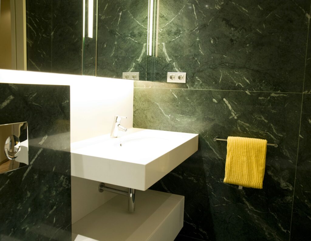 Baño Moderno Elegante con Mármol Verde y Accesorios Blancos Un baño moderno que presenta un elegante lavabo blanco con un grifo contemporáneo sobre un armario minimalista blanco, rodeado de ricas paredes de mármol verde. El espacio está suavemente iluminado desde arriba, creando una atmósfera acogedora. Una toalla amarilla brillante cuelga de un toallero de cromo, añadiendo un vibrante toque de color contra el elegante fondo de piedra.