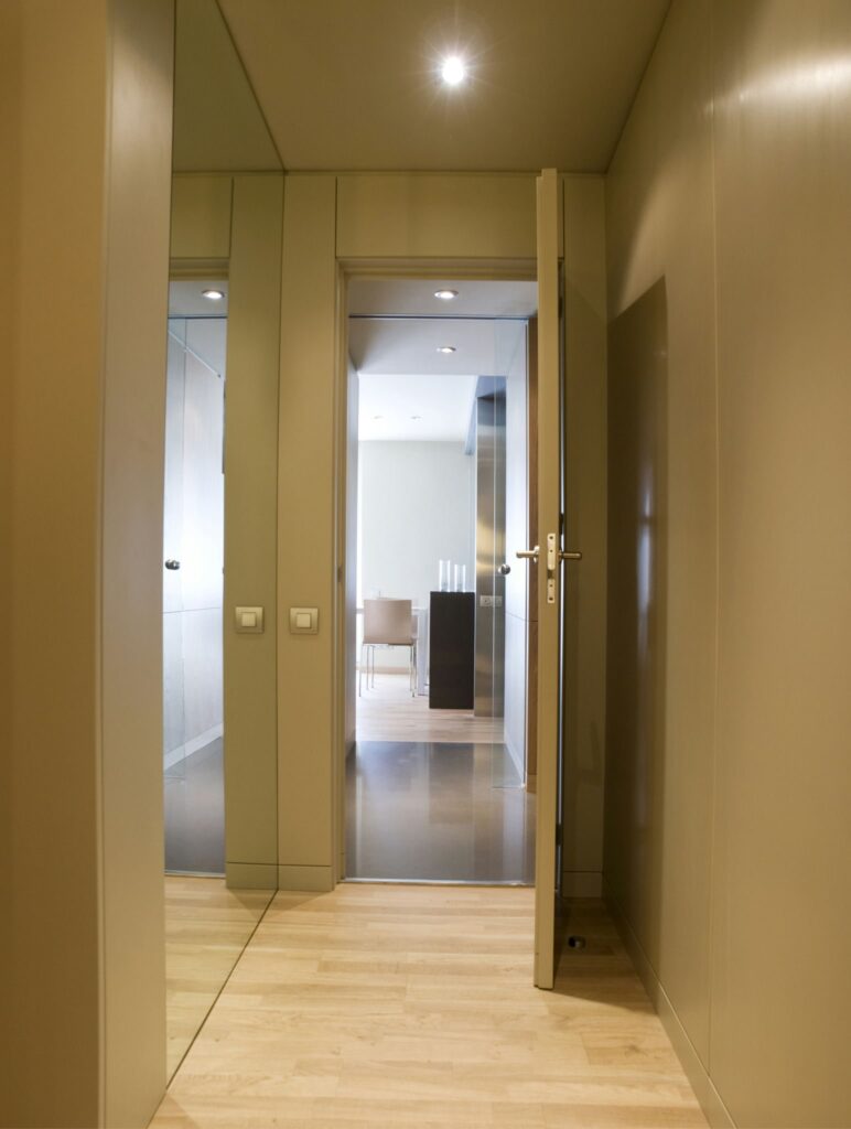 Diseño de pasillo moderno minimalista con concepto de puerta abierta Un pasillo moderno y espacioso con dos puertas en lados opuestos, pintado en tonos neutros. El suelo de madera clara y la iluminación empotrada en el techo crean una atmósfera brillante, mientras que una puerta abierta revela un sencillo comedor con una mesa y sillas.