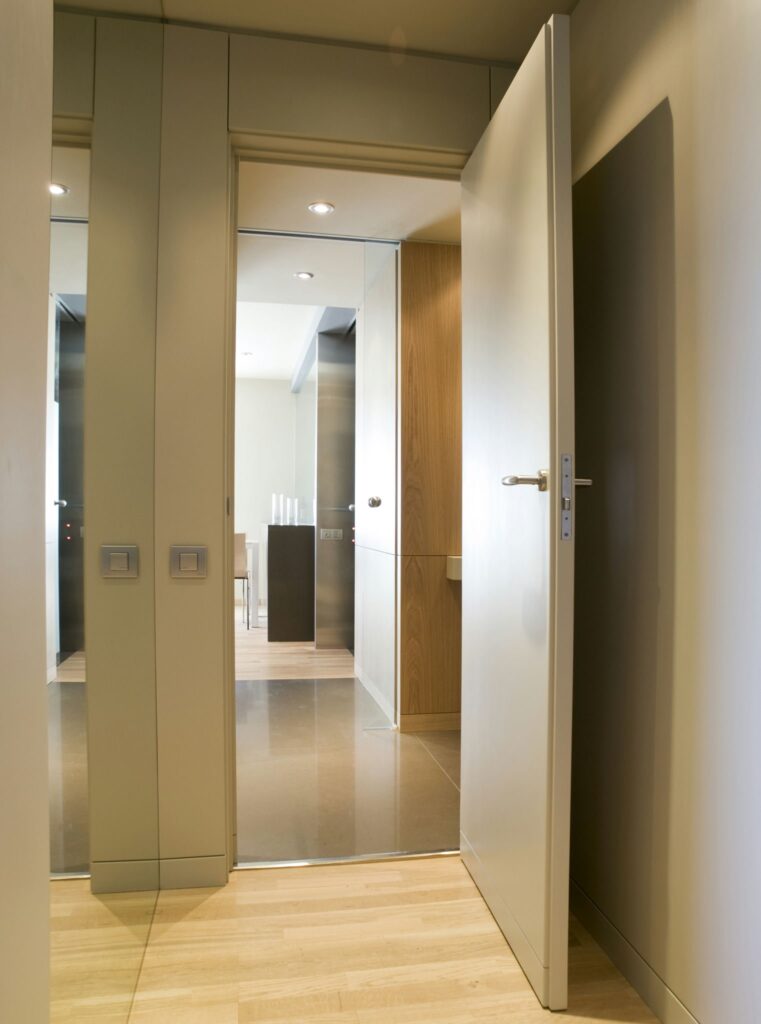 Pasillo interior moderno con puerta abierta hacia el área de comedor contemporáneo Un pasillo interior luminoso y espacioso con paredes de colores claros y pisos de madera clara. Una puerta abierta conduce a una habitación bien iluminada con muebles de comedor informales y gabinetes elegantes, mostrando una estética de diseño moderno. Los interruptores de luz son visibles en el lado izquierdo y se pueden ver reflejos en el piso brillante.