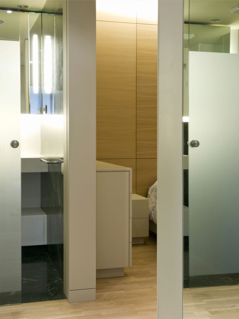 Diseño de Interior Moderno Minimalista con Puertas de Vidrio y Accesorios Elegantes Un interior moderno minimalista que muestra dos puertas de vidrio con manijas de metal circulares. Una puerta se abre a un baño elegante con accesorios elegantes y abundante luz, mientras que la otra conduce a un acogedor dormitorio visible en el fondo. El espacio está complementado con paneles de madera clara y un suelo de madera de color claro, destacando una estética tranquila y contemporánea.