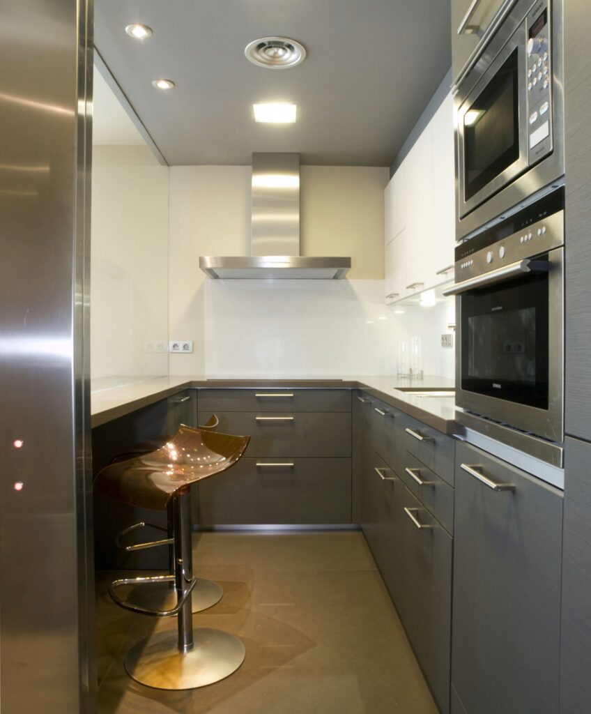 Diseño de Cocina Compacta Moderna con Muebles de Gabinete Gris Oscuro y Electrodomésticos de Acero Inoxidable Un interior de cocina moderna que presenta muebles de gabinete gris oscuro, electrodomésticos de acero inoxidable y una encimera blanca elegante. El diseño incluye un backsplash minimalista, iluminación suave en el techo y grandes azulejos en el suelo, creando una atmósfera acogedora perfecta para comidas informales.