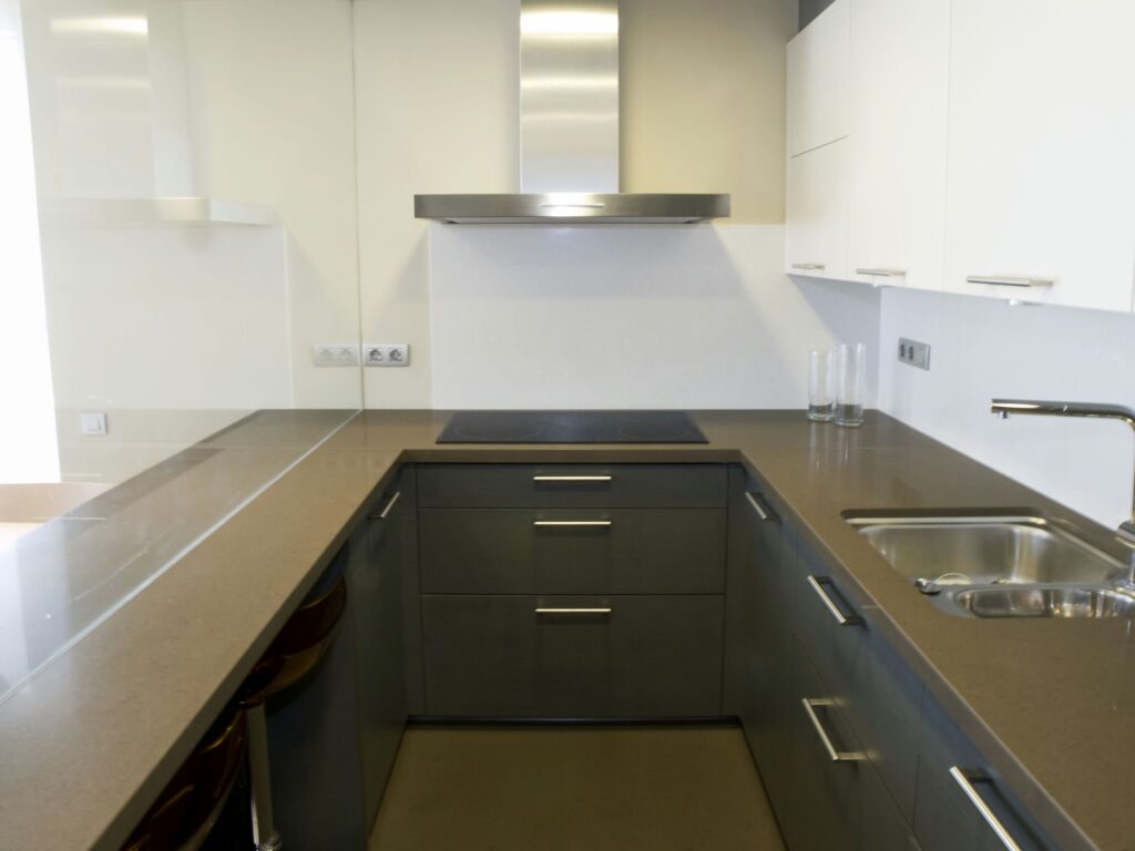 Diseño de Cocina Minimalista Moderna con Muebles Oscuros y Claros Una cocina contemporánea que presenta un diseño elegante, con muebles oscuros, gabinetes superiores de color claro, electrodomésticos de acero inoxidable y una encimera de tono neutro. La luz brillante entra por una ventana, realzando la atmósfera aireada de la habitación.