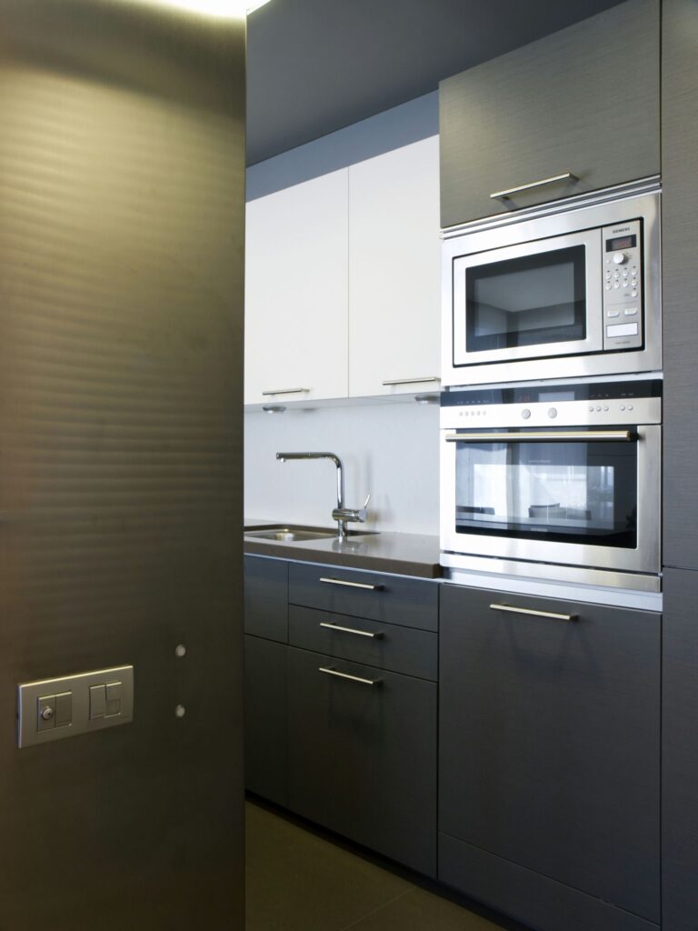 Diseño de cocina moderna minimalista con gabinetes gris oscuro y blancos Una cocina contemporánea que presenta elementos de diseño minimalista, incluyendo gabinetes gris oscuro y blancos, un horno y microondas de acero inoxidable, un fregadero de dos tinas con un grifo plateado, y pintura de pared azul claro.