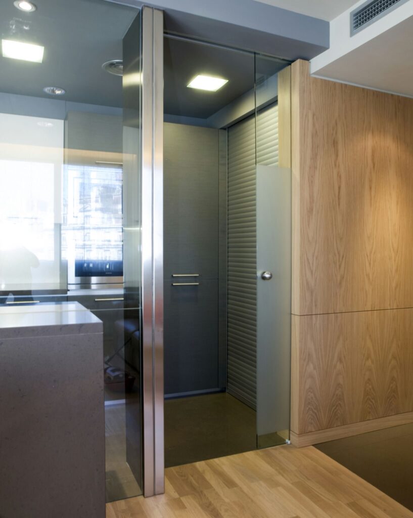 Entrada moderna y elegante con puerta de vidrio y acentos de acero inoxidable Entrada contemporánea con puerta de vidrio, acentos de acero inoxidable, paredes gris oscuro, persiana enrollable texturizada y suelo de madera clara, iluminada por luz empotrada para una estética minimalista.