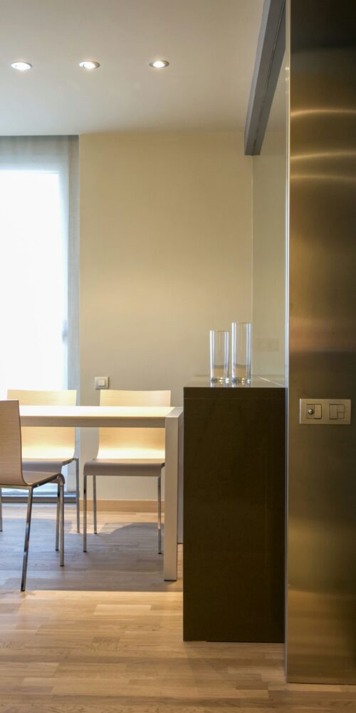 Un área de comedor elegante y minimalista que presenta una mesa cuadrada blanca, sillas de tonos claros y suelo de madera, iluminada por una suave luz natural que entra a través de cortinas ligeras.